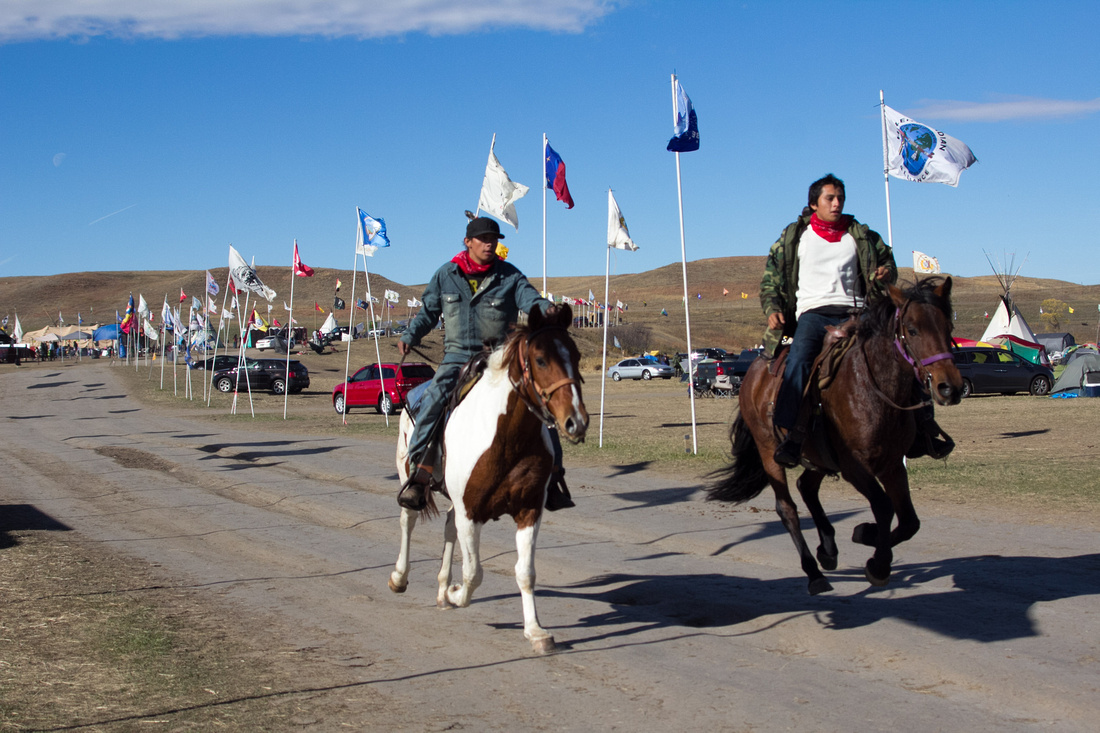 Two Native American men ride their horses through the camp at a quick pace.